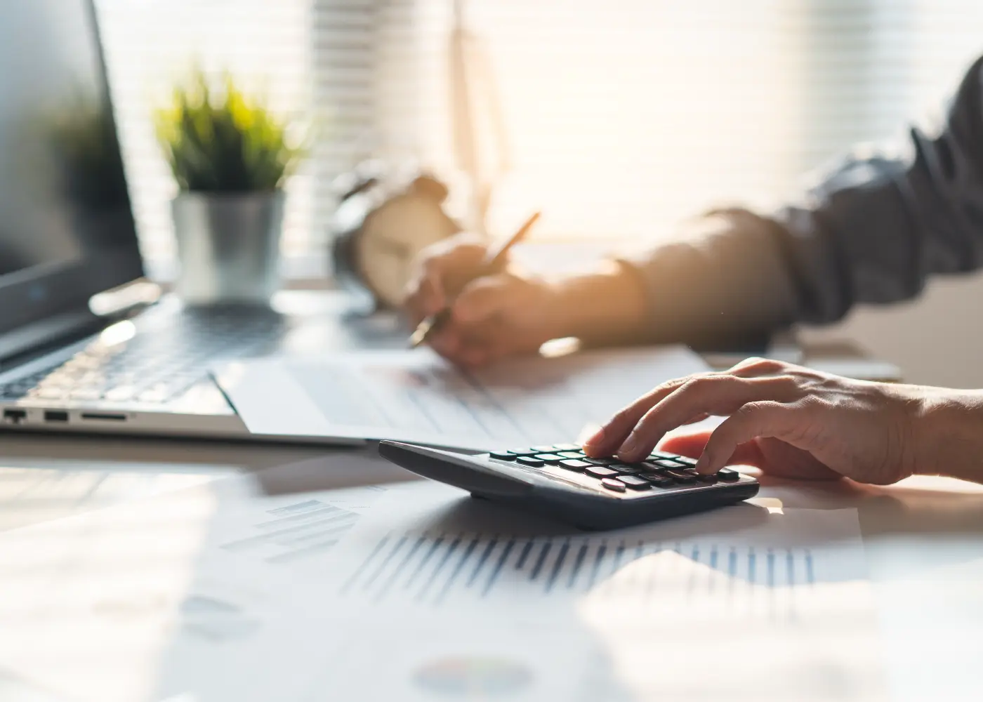 Nahaufnahme eines Schreibtischs mit Laptop, Diagrammen und Dokumenten; im Vordergrund tippt eine Person auf einem Taschenrechner, im Hintergrund notiert sie etwas auf Papier.