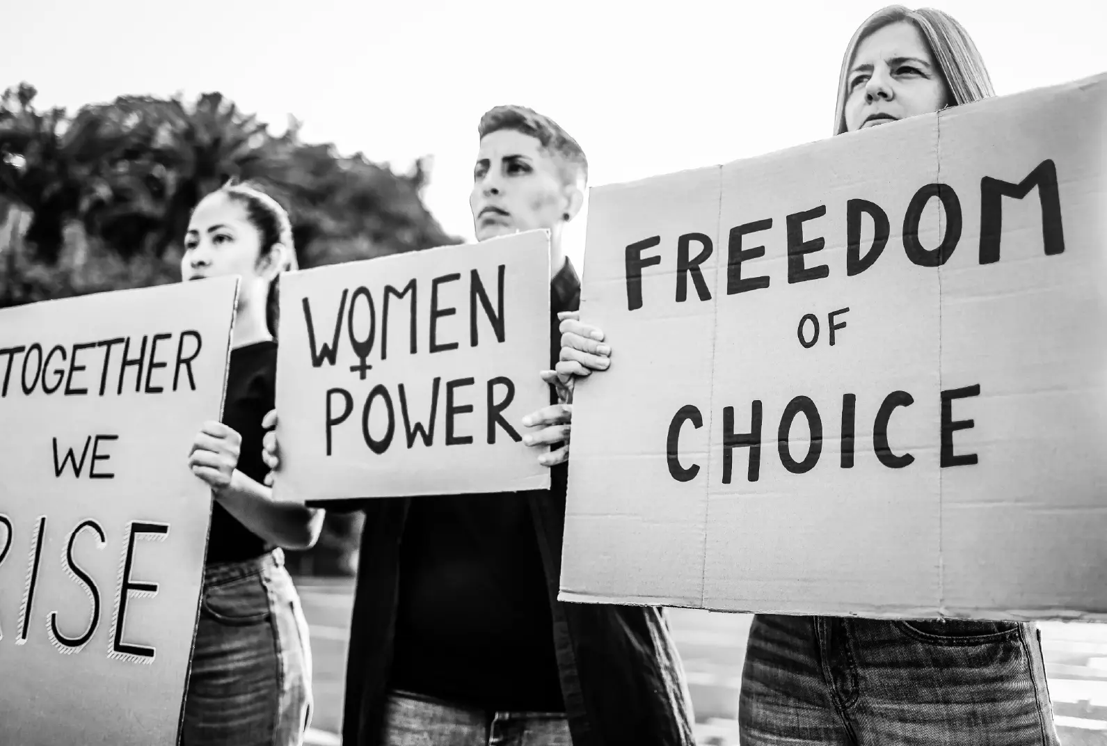 Drei Frauen stehen ernst mit Kartonschildern auf der Straße. Auf den Schildern steht: "Together we rise", "Women Power" und Freedom of choice".